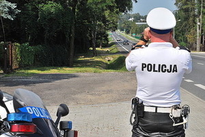 fot. policjant patrzący przez lornetkę, odwrócony tyłem z boku część motocykla policyjnego