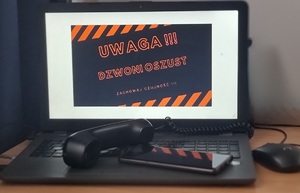 na zdjęciu laptop, na monitorze napis &amp;quot;uwaga dzwoni oszust zachowaj czujność&amp;quot;, na klawiaturze laptopa słuchawka od telefonu i telefon komórkowy